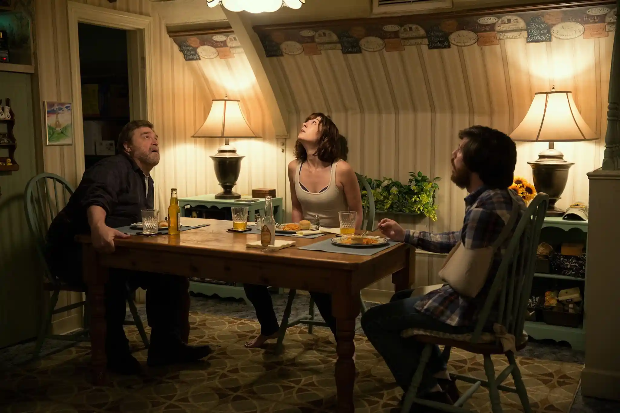 Three people look at the ceiling of an underground bunker, hearing vibrations from outside.